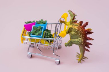 Yumuşak pembe arka planda çiçek saksısı dolu alışveriş arabası olan komik yeşil dinozor oyuncağı.