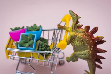 Yumuşak pembe arka planda çiçek saksısı dolu alışveriş arabası olan komik yeşil dinozor oyuncağı.