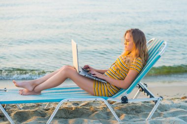 Dizüstü bilgisayar kullanarak deniz görünümü kumsalda şezlong üzerinde oturan genç kadın uygun. Dizüstü bilgisayarda, boş ekran üzerinde sörf kodlama chaise uzunluğundaki salon çalışan kadın serbest programcı. Uzak iş kavramı.