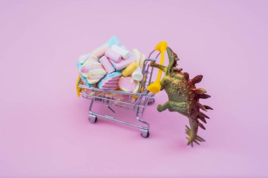 Komik yeşil dinozor oyuncağı. Alışveriş arabası marşmelovla dolu. Yumuşak pembe arka planda.