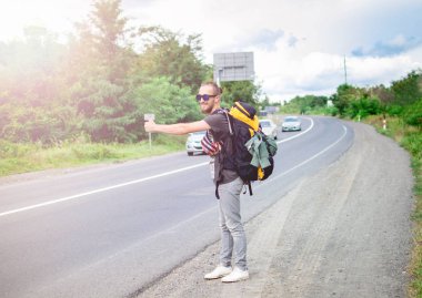 Modayı takip eden genç adam otobanın ortasında dikiliyor ve başka tarafa bakıyor. Sabah yolda yürüyen büyük sırt çantalı bir gezginin tam boy portresi. Maceradaki Duygusal Gezgin.