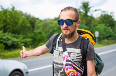 Modayı takip eden genç adam otobanın ortasında dikiliyor ve başka tarafa bakıyor. Sabah yolda yürüyen büyük sırt çantalı bir gezginin tam boy portresi. Maceradaki Duygusal Gezgin.