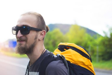 Modayı takip eden genç adam otobanın ortasında dikiliyor ve başka tarafa bakıyor. Sabah yolda yürüyen büyük sırt çantalı bir gezginin tam boy portresi. Maceradaki Duygusal Gezgin.