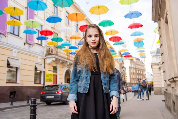 Beautiful girl walking under umbrellas in the city. Cute teenage girl in black dress posing against a background of multicolored umbrellas alley. Spring sunny concept. 8 March. Happy Women\'s Day.