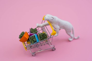 Yumuşak pembe arka planda çiçek saksısı dolu alışveriş arabası olan komik beyaz kaplan oyuncağı.