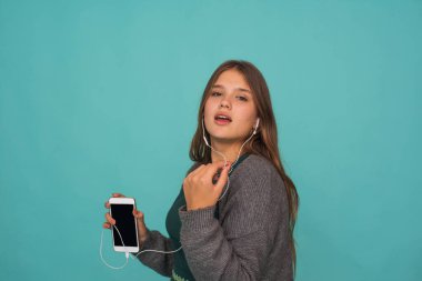Moda oldukça tatlı, kaygısız genç kız kulaklıkla müzik dinliyor ve mavi arka planda akıllı telefonla şarkı söylüyor.