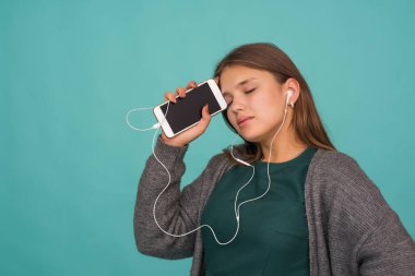Moda oldukça tatlı, kaygısız genç kız kulaklıkla müzik dinliyor ve mavi arka planda akıllı telefonla şarkı söylüyor.