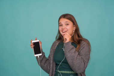 Moda oldukça tatlı kaygısız genç kız kulaklıkla müzik dinliyor, mavi arka planda akıllı telefonla şarkı söylüyor ve konuşuyor.