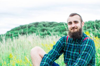 Yakışıklı hippi sakallı, ciddi yüzlü, kumaş gömlekli, askılı, açık havada, dağın tepesinde, bulutlu gökyüzüne karşı. Turizm konsepti. Muhteşem manzara..