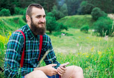 Yakışıklı hippi sakallı, ciddi yüzlü, kumaş gömlekli, askılı, açık havada, dağın tepesinde, bulutlu gökyüzüne karşı. Turizm konsepti. Muhteşem manzara..
