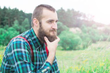 Yakışıklı hippi sakallı, ciddi yüzlü, kumaş gömlekli, askılı, açık havada, dağın tepesinde, bulutlu gökyüzüne karşı. Turizm konsepti. Muhteşem manzara..