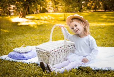 Küçük güzel bir kız piknik sepeti ve hasır şapkayla parkta yürüyor. Komik duygusal bebek.