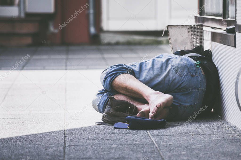 Homeless or drunk man sleeps on the street in the shadow of the building barefooted 