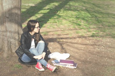 Güzel genç okul veya üniversitede kızla uzun saç, gözlük ve yere kitap ve eğitim sınavı, eğitim kavramı için okuma parkta oturan siyah deri ceket