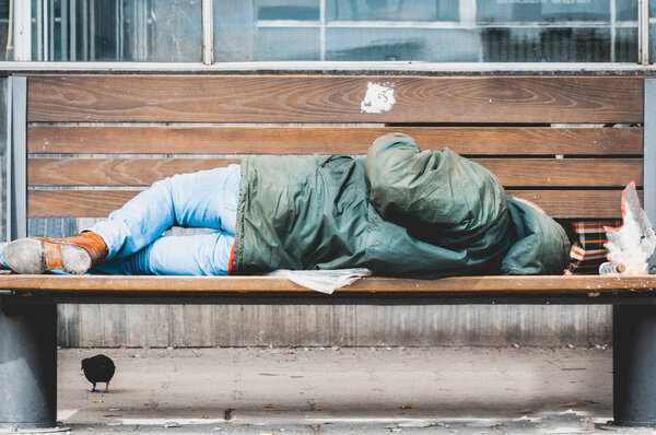 Poor homeless man or refugee sleeping on the wooden bench on the urban street in the city, social documentary concept