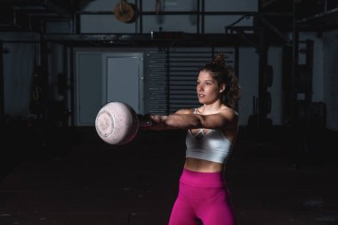 Genç, terli, güçlü, kaslı, formda bir kız elleriyle ağır bir kettlebell tutuyor ve spor salonunda sıkı bir çapraz antrenman yapıyor.