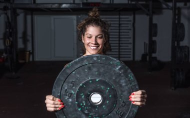 Genç, terli, kaslı, formda, güçlü, büyük halterli bir kız, ağır bir halter tabakasını elleriyle tutuyor ve kaldırıyor. Spor salonunda omuz kasları için antrenman yapıyor. Gerçek insanlar.