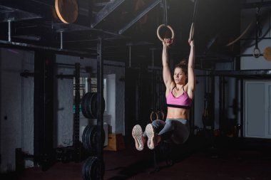 Genç, aktif, güçlü, terli, kaslı, iri kaslı bir kız. Jimnastik halkalarında egzersiz yapıyor. Karın kasları için yüzükler yapıyor.