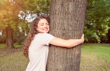 Güzel, gülümseyen, pozitif ve mutlu çevreci bir kız parkta büyük bir ağaca sarılıyor doğayı seviyor ve çevresel konsepti korumak istiyor. Ormanı kurtarmak istiyor.