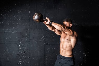 Genç, güçlü, terli, formda, iri kaslı bir adam salıncakta sallanmak için ağır bir kettlebell tutuyor. Spor salonunda sıkı antrenman yapıyor. 