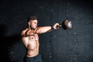 Genç, güçlü, terli, formda, iri kaslı bir adam salıncakta sallanmak için ağır bir kettlebell tutuyor. Spor salonunda sıkı antrenman yapıyor.