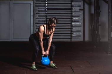 Genç, terli, güçlü, kaslı, formda, elinde ağır bir kettlebell tutan kız, salıncağa hazırlanıyor. Gerçek insanlar spor salonunda sıkı bir çapraz antrenman yapıyor.