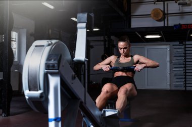Genç, güçlü, güçlü, terli, çekici, kaslı, iri kaslı bir kadın. Spor salonunda kürek çekerken ağır çapraz antrenman yapıyor. Gerçek insanlar kürek çekiyor.