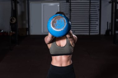 Genç, terli, güçlü, kaslı, formda, elinde ağır bir kettlebell tutan, salıncakta sallanan, sıkı bir çapraz antrenman yapan bir kız. Spor salonunda gerçek insanlar seçici odaklanma bulanıklığı yaşıyor.