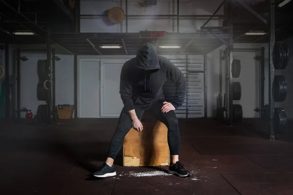 Young fit man in black sweatsuit with hood on his head sitting on the jump box in the gym taking a rest after hard core cross workout training