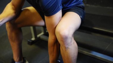 Genç, kaslı, formda bir adam spor salonunda oturuyor ve kaslarını halterle şişiriyor.