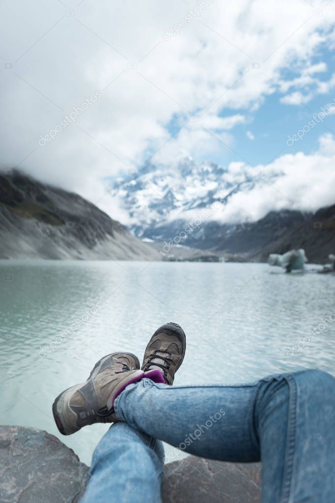 View of woman legs in trekking boots on background with green river, mountains and cloudy sky