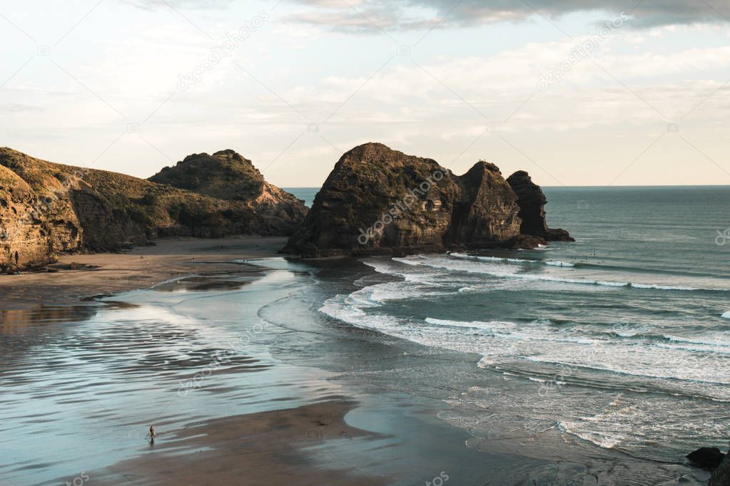 Rock formations on coast of New Zealand Piha beach, gold light in sunset, waves coming to beach.