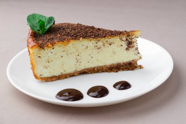 Bej bir masa örtüsünün arka planında beyaz bir tabakta nane yapraklı bir parça cheesecake.