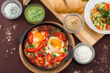 İsrail yumurtalı shake, domatesler ve yeşillikler, siyah dökme demir bir tavada, ahşap bir tahtada ekmek ve kahverengi bir tekstil arka planında humus.