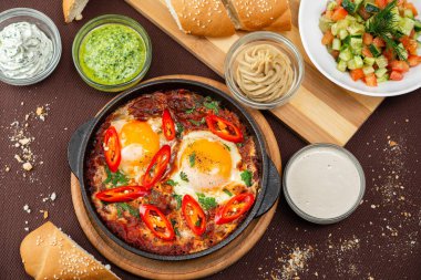 İsrail yumurtalı shake, domatesler ve yeşillikler, siyah dökme demir bir tavada, ahşap bir tahtada ekmek ve kahverengi bir tekstil arka planında humus.