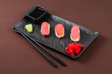 Ton balığı, zencefil, wasabi, soya sosu ve yemek çubuklarıyla suşi, kahverengi tekstil arka planında siyah bir tabakta.