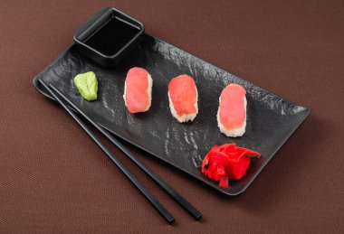 Suşi, somon, zencefil, wasabi, soya sosu ve yemek çubuklarıyla birlikte saşimi. Kahverengi bir tekstil arka planında siyah bir tabakta suşi.