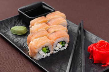 Suşi, somon, ton balığı, pirinç, zencefil, soya sosu ve yemek çubuklarıyla birlikte, siyah bir tabakta, kahverengi bir tekstil arka planında.