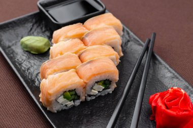 Suşi, somon, ton balığı, pirinç, zencefil, soya sosu ve yemek çubuklarıyla birlikte, siyah bir tabakta, kahverengi bir tekstil arka planında.