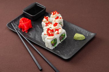 Suşi, somon, ton balığı, pirinç, zencefil, soya sosu ve yemek çubuklarıyla birlikte, siyah bir tabakta, kahverengi bir tekstil arka planında.