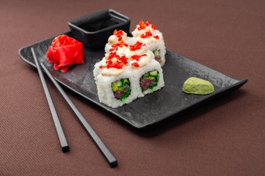 Suşi, somon, ton balığı, pirinç, zencefil, soya sosu ve yemek çubuklarıyla birlikte, siyah bir tabakta, kahverengi bir tekstil arka planında.