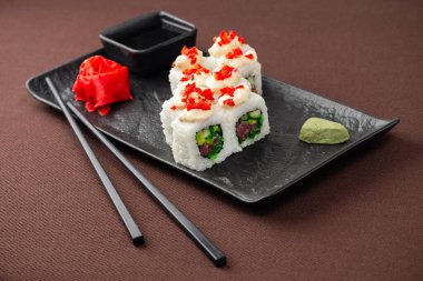 Suşi, somon, ton balığı, pirinç, zencefil, soya sosu ve yemek çubuklarıyla birlikte, siyah bir tabakta, kahverengi bir tekstil arka planında.