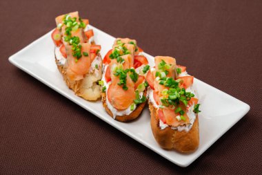 Somon peynirli, domatesli ve soğanlı ekmek sandviçi kahverengi bir tekstil masa örtüsünün üzerinde dikdörtgen beyaz bir tabakta.