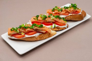 Somon peynirli, domatesli ve soğanlı ekmek sandviçi bej renkli tekstil masa örtüsü üzerinde dikdörtgen beyaz bir tabakta.