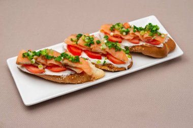 Somon peynirli, domatesli ve soğanlı ekmek sandviçi bej renkli tekstil masa örtüsü üzerinde dikdörtgen beyaz bir tabakta.