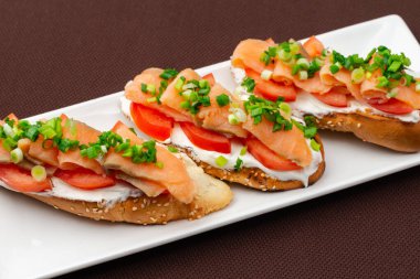 Somon peynirli, domatesli ve soğanlı ekmek sandviçi kahverengi bir tekstil masa örtüsünün üzerinde dikdörtgen beyaz bir tabakta.