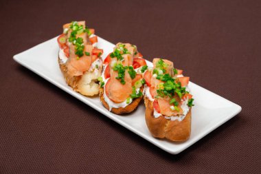 Somon peynirli, domatesli ve soğanlı ekmek sandviçi kahverengi bir tekstil masa örtüsünün üzerinde dikdörtgen beyaz bir tabakta.