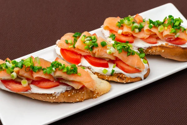 bread sandwich with salmon, cheese, tomatoes and onion on a rectangular white plate on a brown textile tablecloth