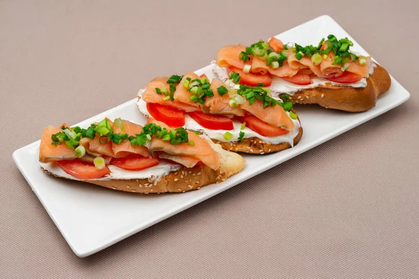bread sandwich with salmon, cheese, tomatoes and onion on a rectangular white plate on a beige textile tablecloth