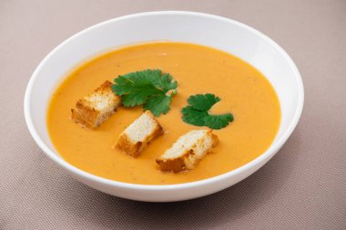 turuncu kremalı balkabağı çorbası, kıtır ekmek ve bej arka planda beyaz bir tabakta otlar.